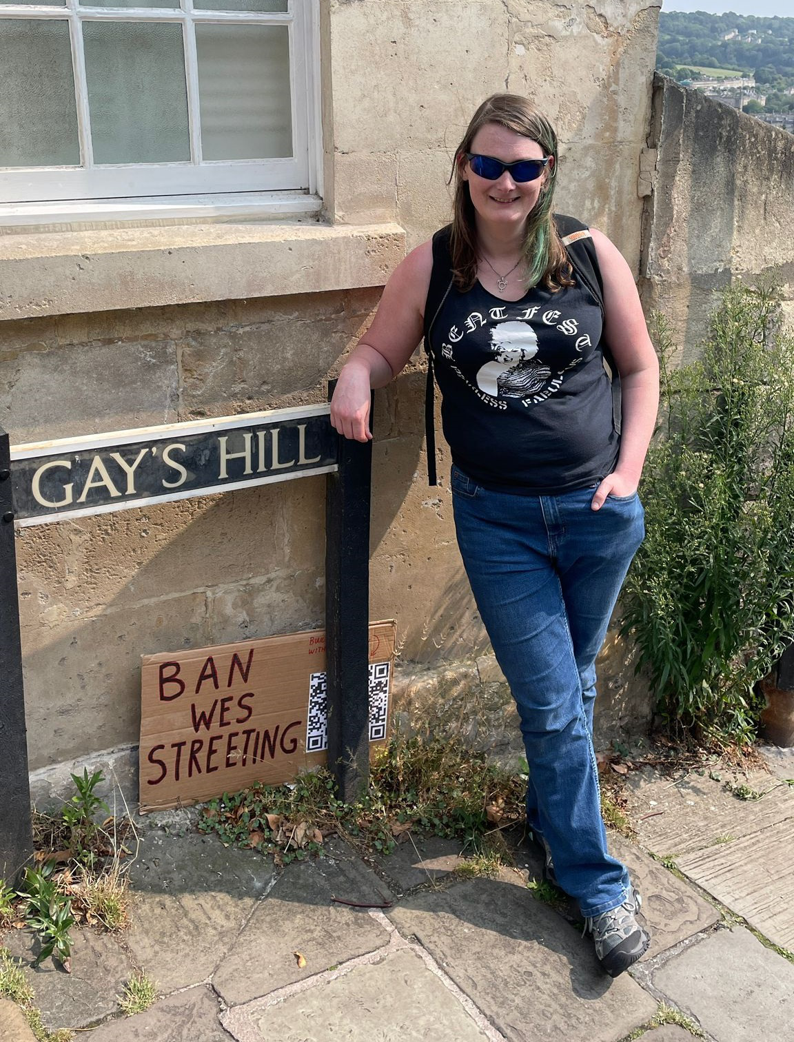 Photo of Ruth posing with a street sign for Gay's Hill, and a placard reading Ban Wes Streeting.
